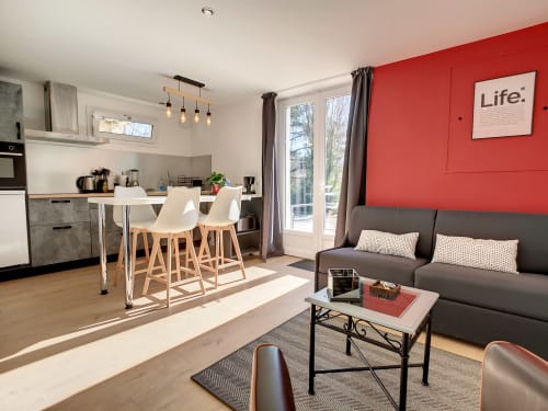 Appartement T1 moderne à la Résidence L'Hermitière avec cuisine équipée, salle à manger, canapé gris et mur d'accent rouge donnant sur jardin