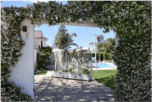 Ingang van Villa Blanche Holidays in Marsala met bloeiende pergola, witte architectuur en zwembadgebied zichtbaar op de achtergrond