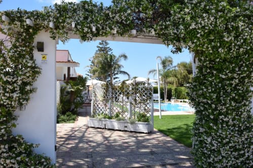 Ingresso ad arco fiorito verso l'area piscina di Villa Blanche Holidays a Marsala con edifici bianchi e palme