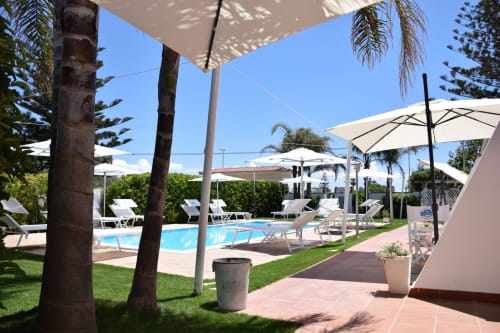 Area piscina Villa Blanche Holidays con palme, ombrelloni bianchi, lettini e vista sul giardino a Marsala, Italia