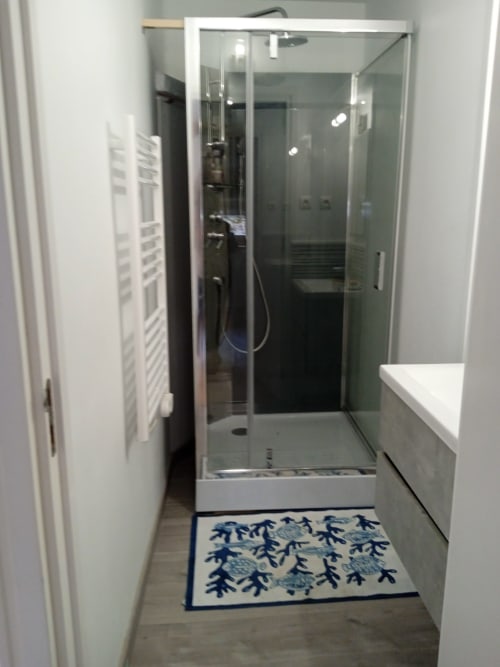 Salle de bain moderne avec cabine de douche en verre, vanité blanche et tapis de bain à motifs bleus à l'hôtel Les Clématites