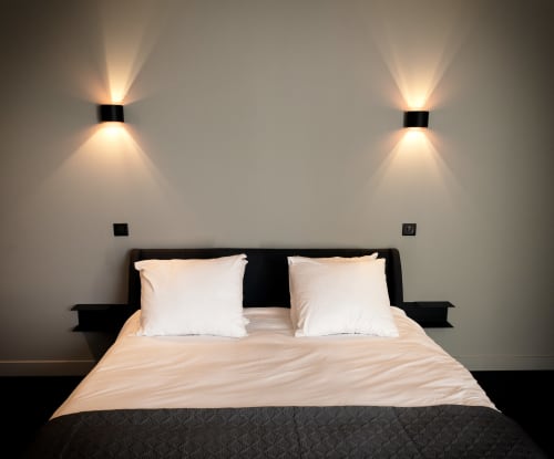 Modern bedroom at Le Petit Château with black bed frame, white pillows, beige bedding, and uplighting on grey walls, Baye,