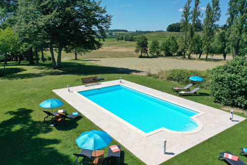 Piscine rectangulaire du Manoir Le Coutureau avec parasols bleus et transats, entourée de verdure et vue sur la campagne