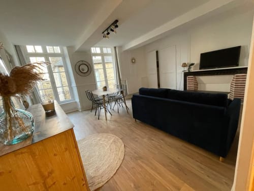 Séjour moderne à Dinan avec canapé noir, TV écran plat, coin repas et vue sur la ville depuis grandes baies