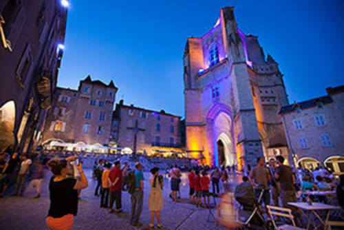 La place historique de Villefranche-de-Rouergue au crépuscule, tour d'église illuminée et foule rassemblée à La Maison De Siloe