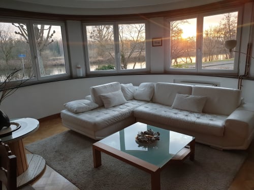Salon lumineux avec canapé-section crème, table basse en verre et baie vitrée panoramique donnant sur la rivière à La Tour De
