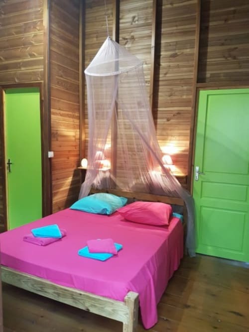 Chambre de villa tropicale avec literie rose, moustiquaire en dais, coussins turquoise, murs en bois et portes vertes