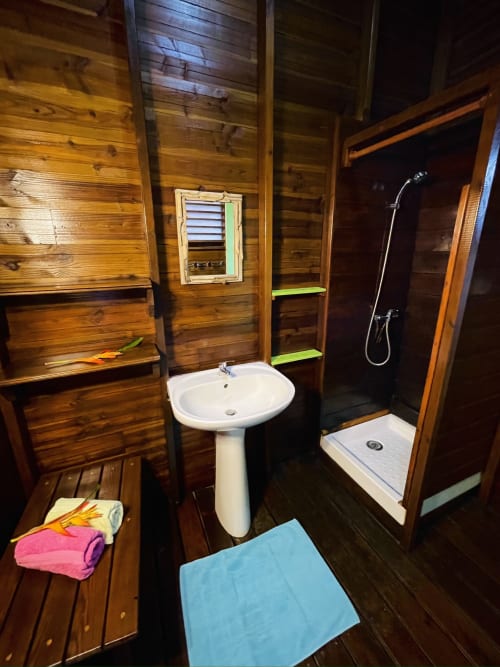 Salle de bain en bois de la villa Les Hauts De Deshaies avec lavabo sur colonne, douche et tapis turquoise