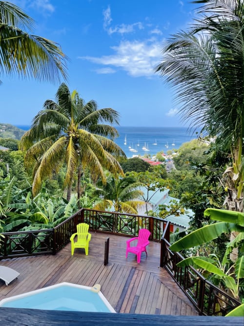 Terrasse de villa tropicale aux Hauts de Deshaies avec chaises colorées, piscine, palmiers et vue sur l'océan
