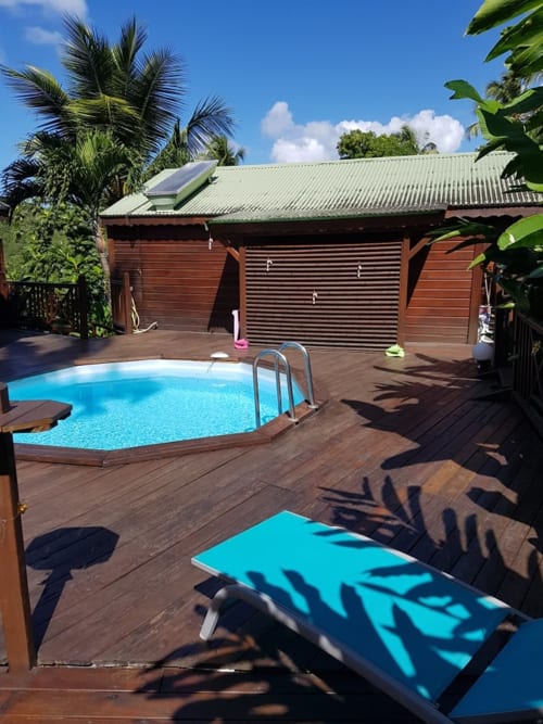 Jardin tropical avec piscine, terrasse en bois, palmiers et petit gîte rouge-brun aux Hauts de Deshaies, Guadeloupe