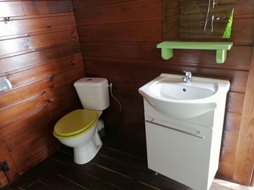 Salle de bain du gîte Les Hauts De Deshaies avec murs en bois, vasque blanche, toilettes avec siège jaune et étagère verte