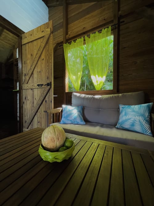 Intérieur de cottage tropical aux Hauts de Deshaies avec terrasse en bois, lit de jour gris, coussins aux motifs de feuilles bleues et rideaux colorés