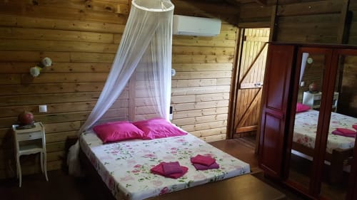 Chambre douillette aux Hauts De Deshaies avec lit à baldaquin, oreillers roses, literie fleurie, murs bois et vue mer