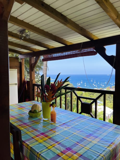 Balcon de cottage face à la mer aux Hauts de Deshaies : vue sur la baie, table colorée, fleurs tropicales et voiliers ancrés.