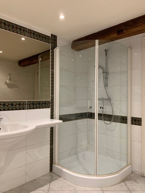 Modern bathroom with curved glass shower enclosure, white tiles, decorative dark border, sink, and rustic wooden beam at Le