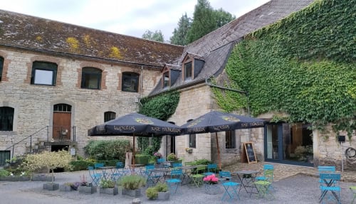 La cour de l'hôtel Le Moulin Des Ramiers à Crupet avec bâtisse en pierre, chaises bleues, parasols et murs couverts de lierre.
