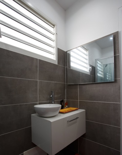 Salle de bain moderne du cottage Kaz A Bibi à Sainte-Rose avec vasque ronde blanche, murs en carrelage foncé et fenêtre à persiennes