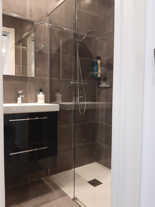Baño moderno en el hostel Room Boutique 32 de Denia con ducha de baldosas oscuras, mamparas de cristal, lavabo blanco y tocador negro
