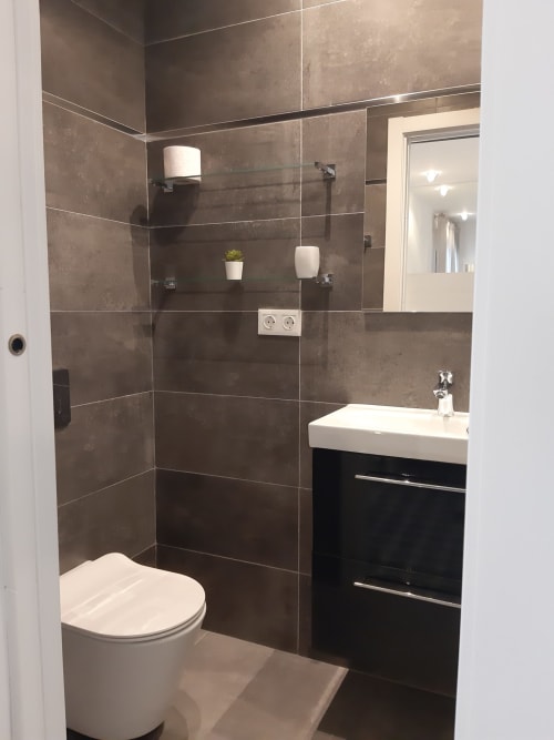 Baño moderno en la hostal Room Boutique 32 de Denia con azulejos oscuros, lavabo blanco, inodoro y ducha