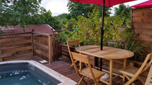 Terrasse avec bain à remous, terrasse en bois, table à manger, parasol rouge et vue sur le jardin luxuriant de la Résidence Les 2 Moulins