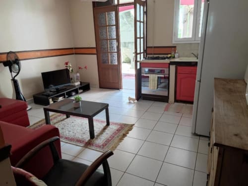 Séjour ouvert à la Résidence Les 2 Moulins avec chaises rouges, table noire, TV, kitchenette et carrelage