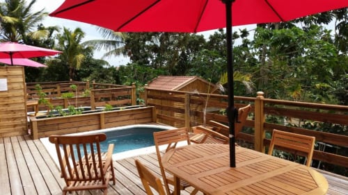 Terrasse tropicale à la Résidence Les 2 Moulins avec vue sur la piscine, terrasse en bois, parasols rouges et jardin luxuriant