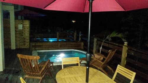 Coin détente au bord de la piscine de nuit à la Résidence Les 2 Moulins avec bain à remous illuminé, chaises en bois sous parasol rose