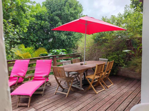 Terrasse colorée de la Résidence Les 2 Moulins avec parasol rouge, chaises longues roses, table à manger en bois et végétation tropicale luxuriante