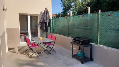 Patio du cottage Chez Anne Marie avec table à manger, chaises rouges, barbecue et clôture de verdure à Beausemblant