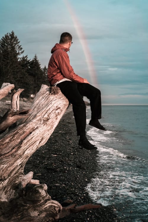 Person in red jacket sitting on driftwood by ocean with rainbow, coastal beach scene near Grignan
