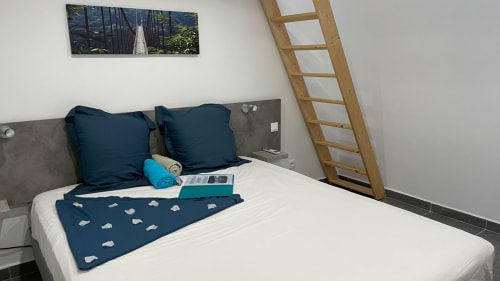 Chambre moderne de la villa Kaz A Bibi avec lit blanc, oreillers bleu marine, échelle en bois et photo de pont au-dessus