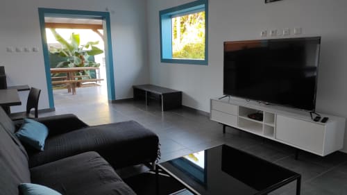 Salon moderne de la villa Kaz A Bibi à Sainte-Rose avec télévision écran plat, canapé noir et portes-fenêtres à cadre bleu