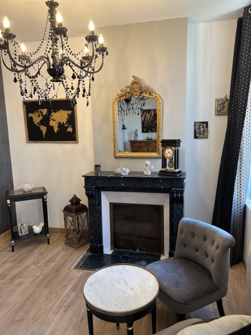 Gabrielle-Chanel-Zimmer des Château Ségot mit Kamin, goldenem Spiegel, schwarzem Kronleuchter und grauem Sessel in Neuilly-le-Réal