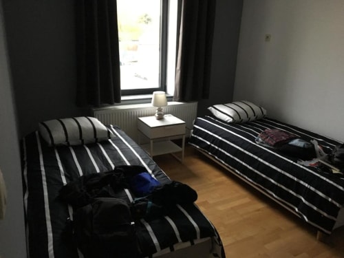 Twin bedroom with striped bedding, small table with lamp, and window with city view at Appartements Courcelles in Belgium