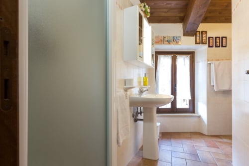 Bagno spazioso al B&B Dormire Assisi con lavabo su colonna, doccia, travi in legno e pavimentazione in cotto