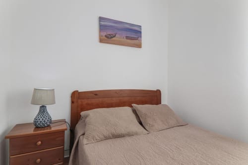 Bedroom at Armação Beach Residences with wooden bed, beige bedding, nightstand with lamp, and coastal artwork above
