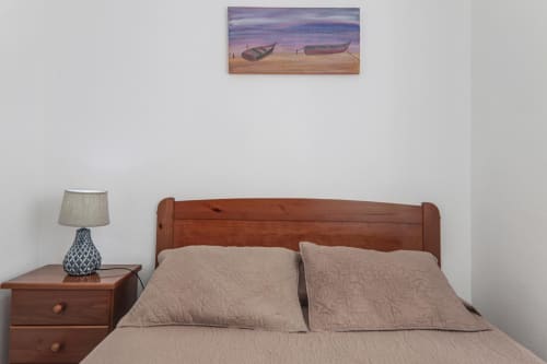 Cozy bedroom at Armação Beach Residences with wooden bed, beige pillows, bedside lamp, and beach boat artwork above headboard