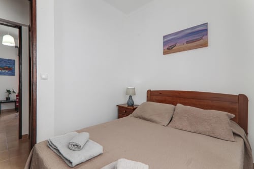 Cozy bedroom at Armação Beach Residences with wooden bed, beige linens, bedside lamp, and seascape artwork in Armação de