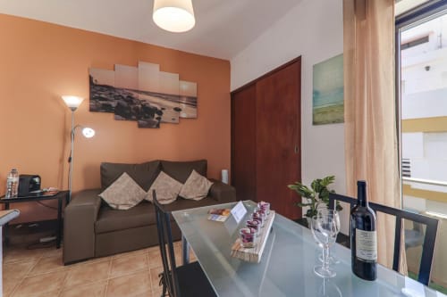 Modern T1 apartment suite at Armação Beach Residences with orange accent wall, brown sofa, dining area, and city view balcony