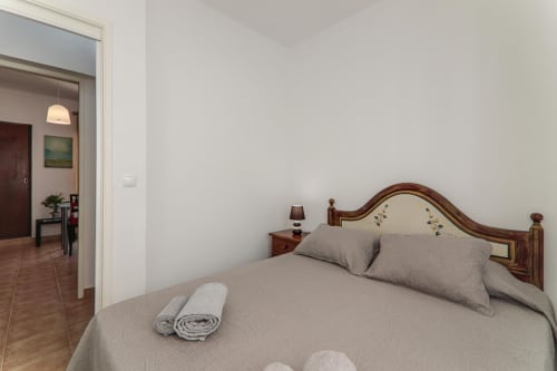 Cozy bedroom at Armação Beach Residences with wooden bed frame, gray bedding, nightstand lamp, and open doorway to living