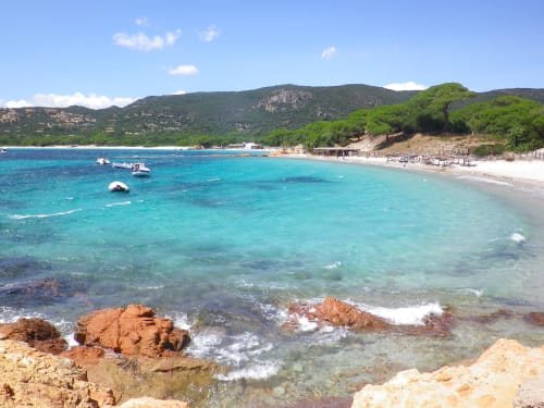 Plage méditerranéenne turquoise à Corbara, Corse avec côte rocheuse, bateaux et montagnes boisées