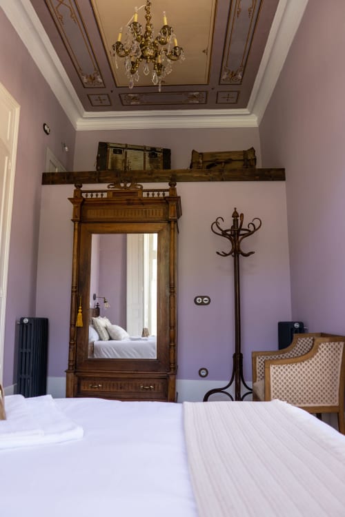 Elegante dormitorio de La Venta de Miyares con paredes lavanda, espejo de madera ornamental, lámpara de cristal y muebles de época