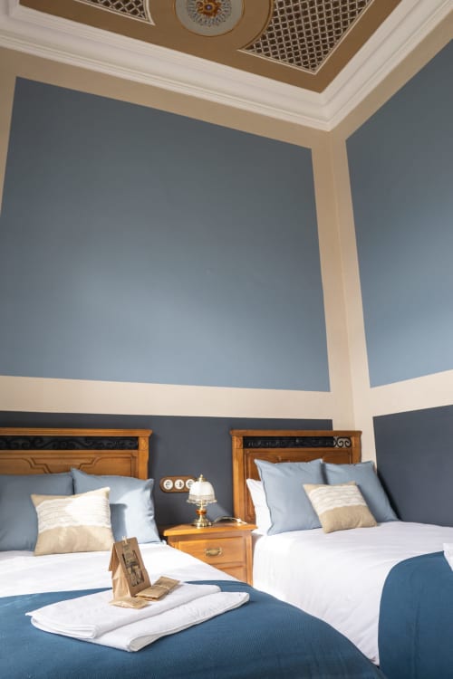 Elegante habitación de hotel con dos camas de madera, paredes azules, detalles de techo blanco y cojines decorativos en La Venta de Miyares.