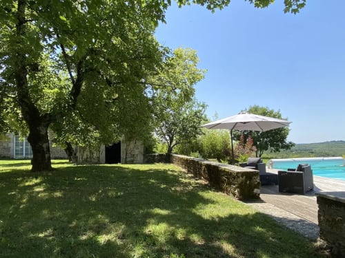 Jardin piscine des Lumieres Du Causse avec bâtisse en pierre, arbres centenaires, salon sous parasol et piscine turquoise