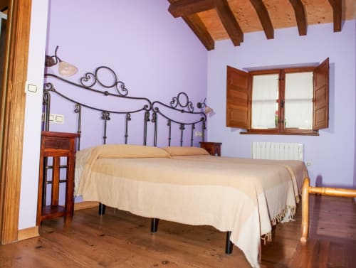 Amplio dormitorio con paredes lavanda, vigas de madera, cama de hierro forjado ornamental y ropa de cama crema en Turismo en Sanabria