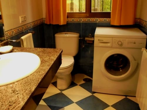 Banheiro do hotel Sanabria com pia, vaso sanitário, máquina de lavar, piso de azulejos em padrão losango preto e bege, e cortinas laranja