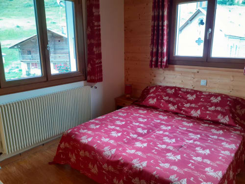Chambre lumineuse avec vue sur les montagnes au Chalet Les Arcosses, literie alpine rouge, murs en bois et grandes fenêtres donnant sur La