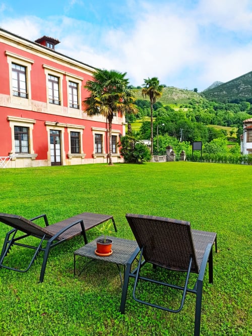 Lounge chairs on green lawn with red colonial building and palm trees at La Venta de Miyares hotel in Asturias
