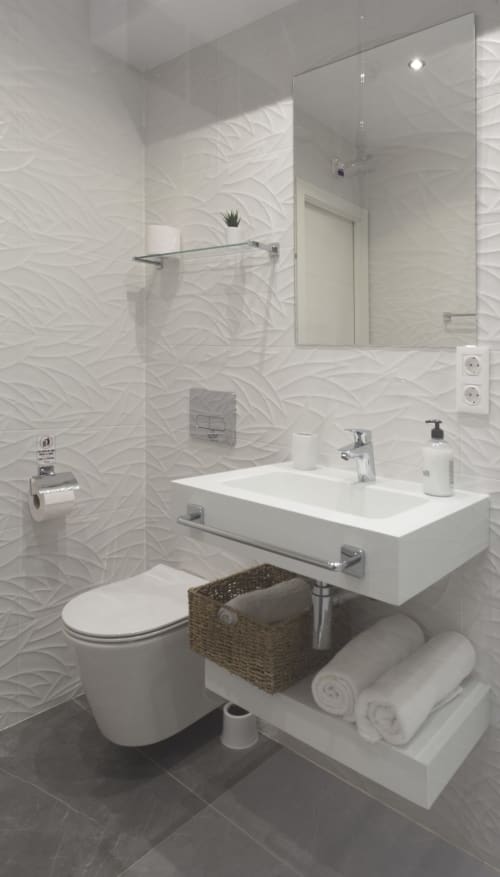 Baño moderno blanco con paredes de azulejos texturados, ducha de vidrio, lavamanos, inodoro y almacenamiento tejido en el hostal Room Boutique 32