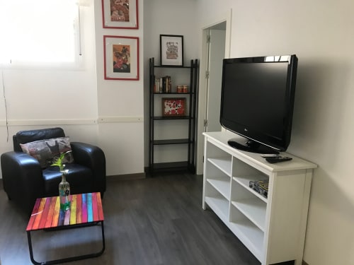 Elegante sala de estar con sillón de cuero negro, mesa de café colorida, televisor en soporte blanco y estantes en Apartamentos